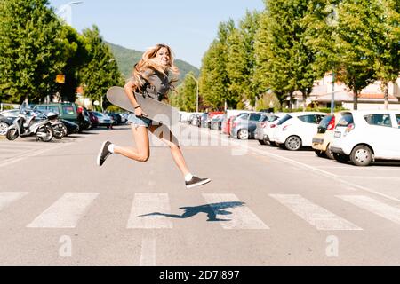 Fröhliche junge Frau hält Skateboard beim Springen auf Zebra Kreuzung In der Stadt Stockfoto