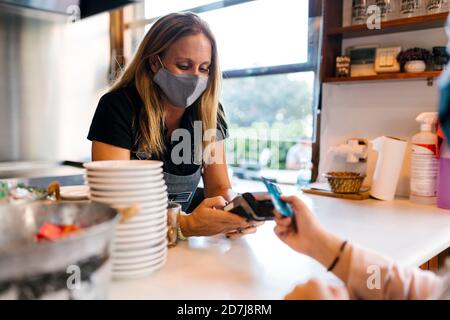 Weibliche Kassiererin in Gesichtsmaske, die die Zahlung per Kreditkarte erhält Im Café während der COVID-19 Stockfoto