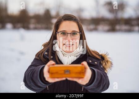 Nahaufnahme einer lächelnden jungen Frau, die im Stehen ein Weihnachtsgeschenk hält Im Winter im Freien Stockfoto