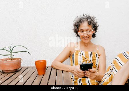 Lächelnde reife Frau mit kurzen Haaren mit Smartphone während Sitzen gegen weiße Wand im Hinterhof Stockfoto