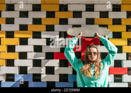 Junge Frau hebt Skateboard, während sie gegen Backsteinwand steht Stockfoto