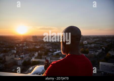 Glatze Mann mit Smartphone für das Fotografieren Sonnenuntergang über der Stadt Von der Terrasse Stockfoto