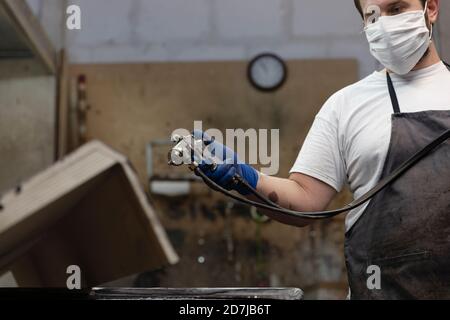 Mann trägt Gesichtsmaske mit Spritzpistole auf Holz Während des Standens in der Fabrik Stockfoto