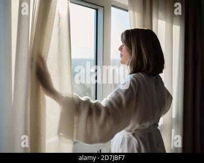 Blonde ältere Frau öffnet weißen Vorhang beim Blick durch Fenster In einem luxuriösen Hotelzimmer Stockfoto
