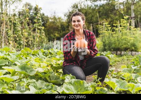 Lächelnde junge Frau, die Squash hält, während sie inmitten von Pflanzen kniet Gemüsegarten Stockfoto