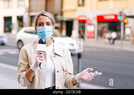 Reporterin trägt Maske und spricht über das Mikrofon, während sie auf steht Straße in der Stadt Stockfoto