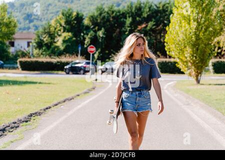 Schöne junge blonde Frau, die weg schaut, während sie Skateboard und hält Wandern auf der Straße in der Stadt während des sonnigen Tages Stockfoto