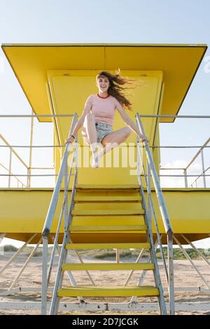 Fröhliche junge Frau, die auf den Stufen der Rettungsschwimmerhütte springt Strand Stockfoto