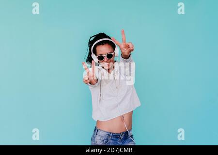 Glückliche modische Frau zeigt Frieden Zeichen beim Hören von Musik durch Kopfhörer vor türkisfarbenem Hintergrund Stockfoto
