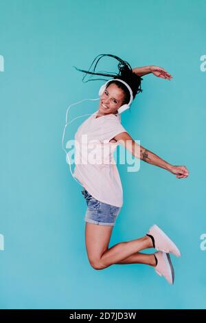 Aufgeregt Frau springen, während Musik durch Kopfhörer gegen türkis hören Hintergrund Stockfoto