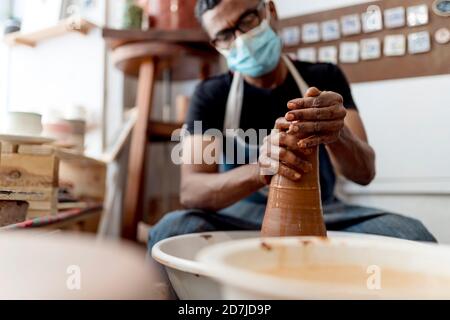 Männliche Töpfer trägt Maske Formform Ton während des Sitzens In der Werkstatt Stockfoto