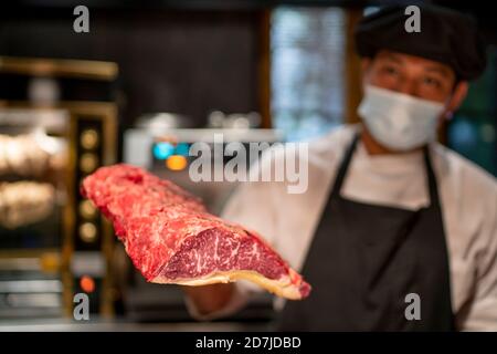 Koch trägt eine schützende Gesichtsmaske und hält Rindfleisch im Restaurant Stockfoto
