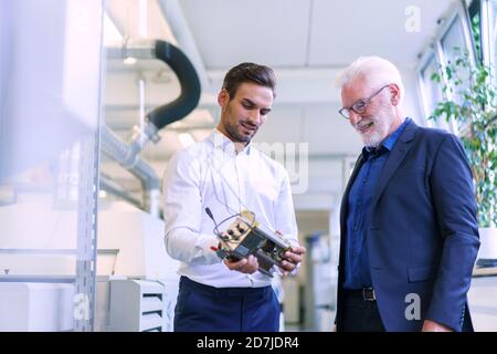 Junger männlicher Ingenieur hält Maschinenteil, während er mit Lächeln diskutiert Leitender Manager im Werk Stockfoto