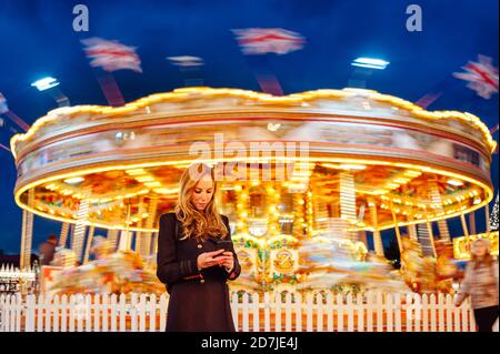 Frau mit Handy gegen beleuchtetes Karussell in der Nacht während Weihnachten Stockfoto