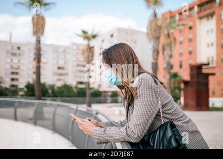 Junge Frau trägt Gesichtsmaske, während mit Smartphone lehnend Auf Geländer in der Stadt Stockfoto