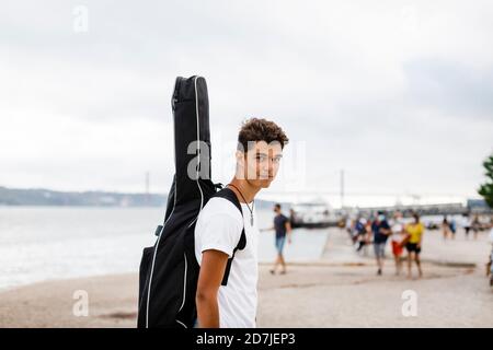 Selbstbewusster, hübscher junger Mann, der Gitarre am Strand gegen den Himmel trägt Stockfoto