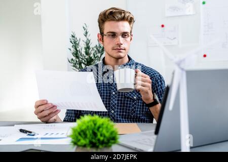 Junger Mann, der beim Kaffeetrinken im Büro auf den Laptop schaut Stockfoto