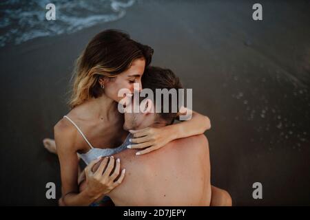 Mann umarmt Frau, während sie am Strand sitzt Stockfoto