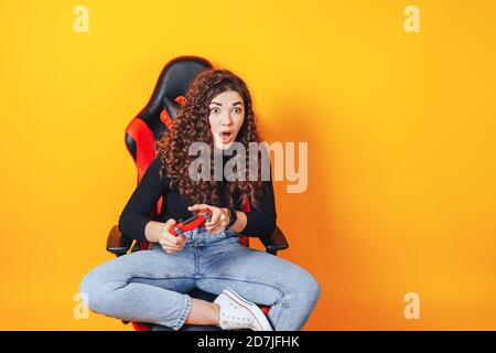 Frau, die hinter einem Spielstuhl in ihren Händen sitzt und rot hält Gamepad auf gelbem Hintergrund Stockfoto