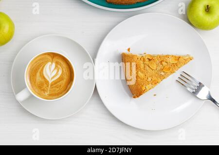 Stück Apfelkuchen mit Mandelblüten und Tasse Kaffee Cappuccino auf weißem Holztisch. Draufsicht. Stockfoto