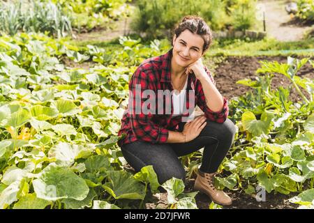 Junge Frau mit der Hand auf dem Kinn knieend inmitten Gemüse wächst Im Garten Stockfoto