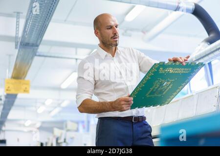 Selbstbewusster Mann-Ingenieur mit Blick auf große Leiterplatte während Untersuchung in beleuchteter Fabrik Stockfoto