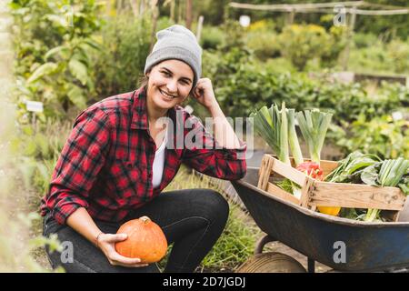 Lächelnde junge Frau hält Squash an Schubkarre im Gemüsegarten Stockfoto
