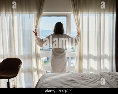 Frau öffnet weißen Vorhang, während Sie durch das Fenster auf Luxus Hotelzimmer Stockfoto