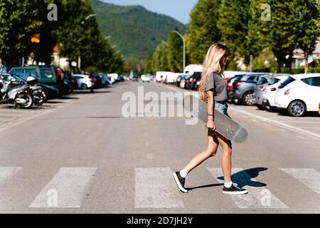 Junge blonde Frau hält Skateboard beim Gehen auf Zebra Kreuzung In der Stadt Stockfoto