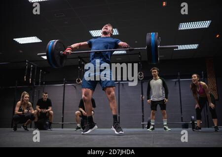 Adaptiver Athlet, der beim Heben der Langhantel mit anfeuernden Menschen springt Hintergrund im Fitnessstudio Stockfoto