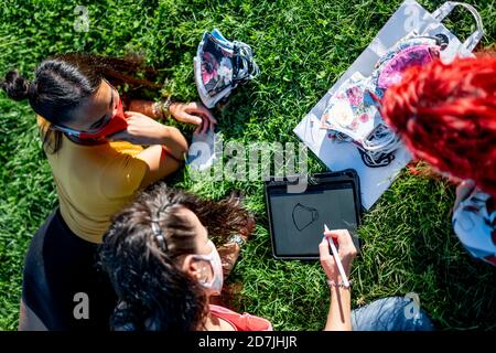 High-Angle-Ansicht von Freunden entwerfen schützende Gesichtsmaske mit Digitales Tablet, während Sie auf dem Rasen sitzen Stockfoto