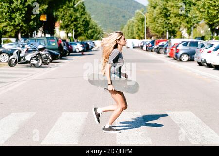 Junge blonde Frau hält Skateboard, während sie auf Zebrastreifen läuft In der Stadt Stockfoto