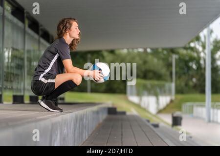 Nachdenkliche Spielerin, die den Fußball hält, während sie auf Stufen hockend Stockfoto