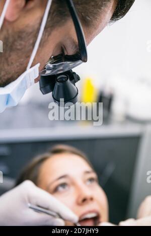 Männlicher Zahnarzt in chirurgischen Lupen, die Zähne von weiblichen Patienten untersuchen In der Klinik Stockfoto