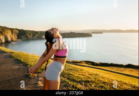 Junge Frau streckt sich die Hände, während sie auf einer Klippe gegen klar steht Himmel bei Sonnenuntergang Stockfoto