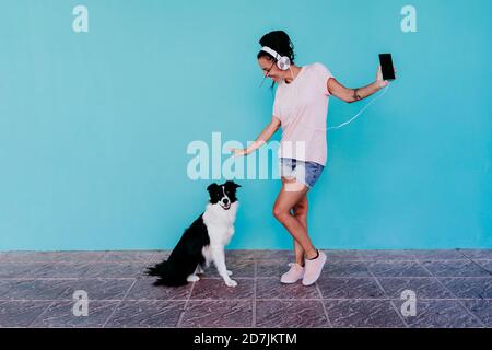 Glückliche Frau, die Musik über Kopfhörer hört, während sie durch Border tanzt Collie Hund gegen türkisfarbene Wand Stockfoto