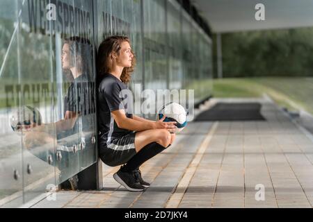 Nachdenkliche Fußballspielerin, die den Ball hält, während sie durch das Glas hocken Wand auf Fußweg Stockfoto