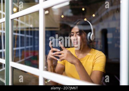 Schöne junge Frau, die Musik hört, während sie Smartphone verwendet Bar durch das Fenster gesehen Stockfoto