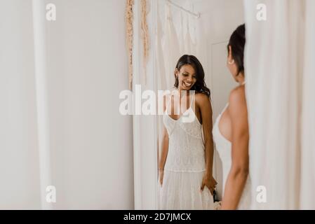 Lächelnde Braut, die im Spiegel schaut, während sie am Hochzeitskleid steht Kaufen Stockfoto