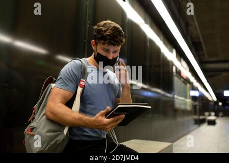 Junger Mann trägt Schutzmaske Gesicht sprechen am Telefon, während Halten Sie während einer Pandemie ein digitales Tablet an der U-Bahn Stockfoto