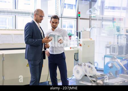 Selbstbewusste männliche Kollegen diskutieren über digitale Tablets, während sie dagegen stehen Maschinen im Werk Stockfoto