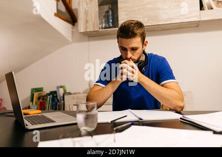 Engagierter junger männlicher Student sitzt mit Händen geklammert während tun Hausaufgaben am Tisch Stockfoto