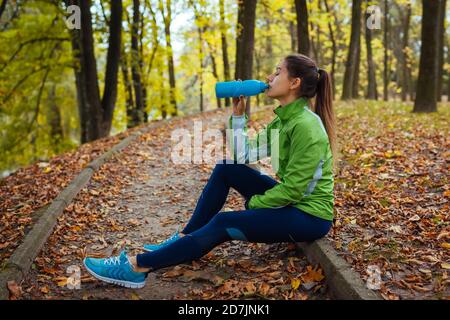 Läufer, der nach dem Training im Herbstpark ausruhend ist. Frau, die Wasser trinkt. Sportlicher, aktiver Lebensstil Stockfoto