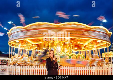 Frau mit Smartphone, während sie gegen beleuchtetes Karussell steht Nacht zu Weihnachten Stockfoto