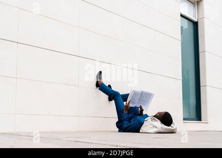 Männlicher Unternehmer liegt mit Seesack auf Bürgersteig beim Lesen Buchen Sie in der Stadt Stockfoto