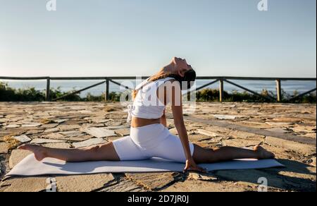 Junge Frau macht Splits auf Trainingsmatte gegen klaren Himmel Bei Sonnenuntergang Stockfoto