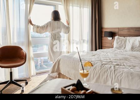 Rentnerin, die den Vorhang öffnete, während sie durch das Fenster schaute Luxuriöses Hotelzimmer Stockfoto