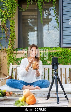 Lächelnd weibliche Lebensmittel-Expertin hält Saft Glas aus dem Glas während Vlogging über Handy im Hinterhof Stockfoto