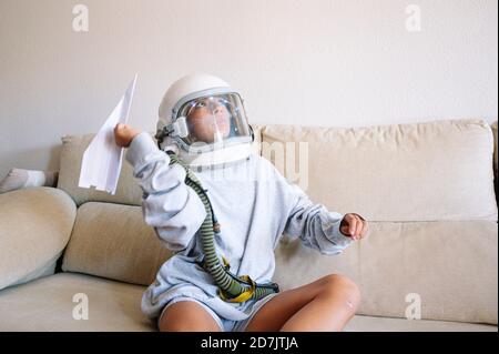 Junge trägt Raum Helm fliegen Papier Flugzeug, während sitzend auf Sofa zu Hause Stockfoto