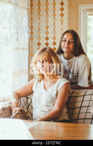 Liebevolle Erwachsene 20s Tochter Umarmung ältere Mutter von hinten während Mutter auf Couch sitzend Menschen posieren Blick auf Kamera lächelnd fühlt sich glücklich, Konzept der mu Stockfoto